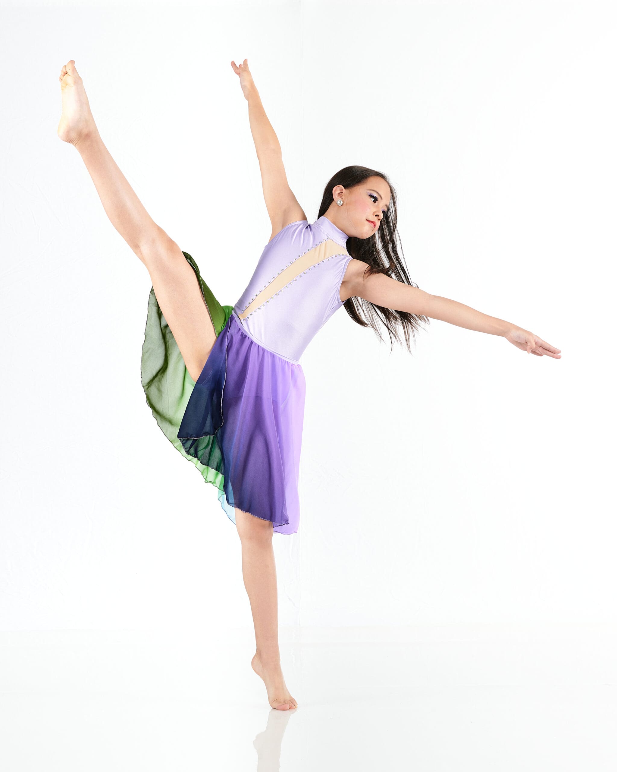 A dancer in a purple and green dress performs a high leg extension with one arm raised and the other extended, balancing gracefully on one foot against a white background.