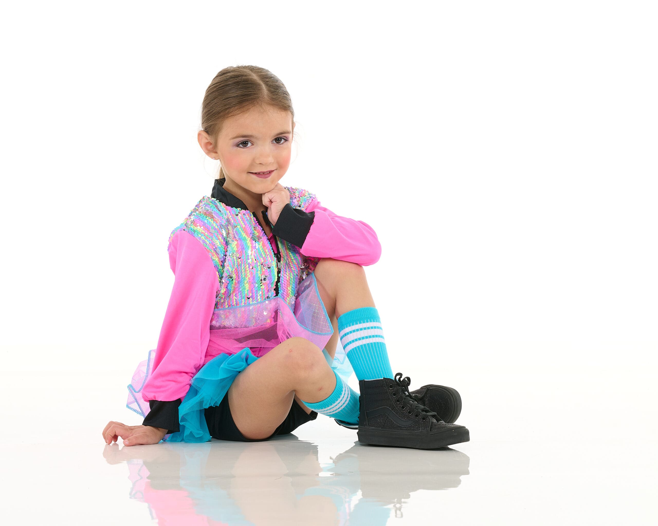 A young girl in a colorful dance costume with a pink top, blue tutu, and striped knee socks sits on the floor with one leg bent and her hand resting on her chin, smiling at the camera against a white background.