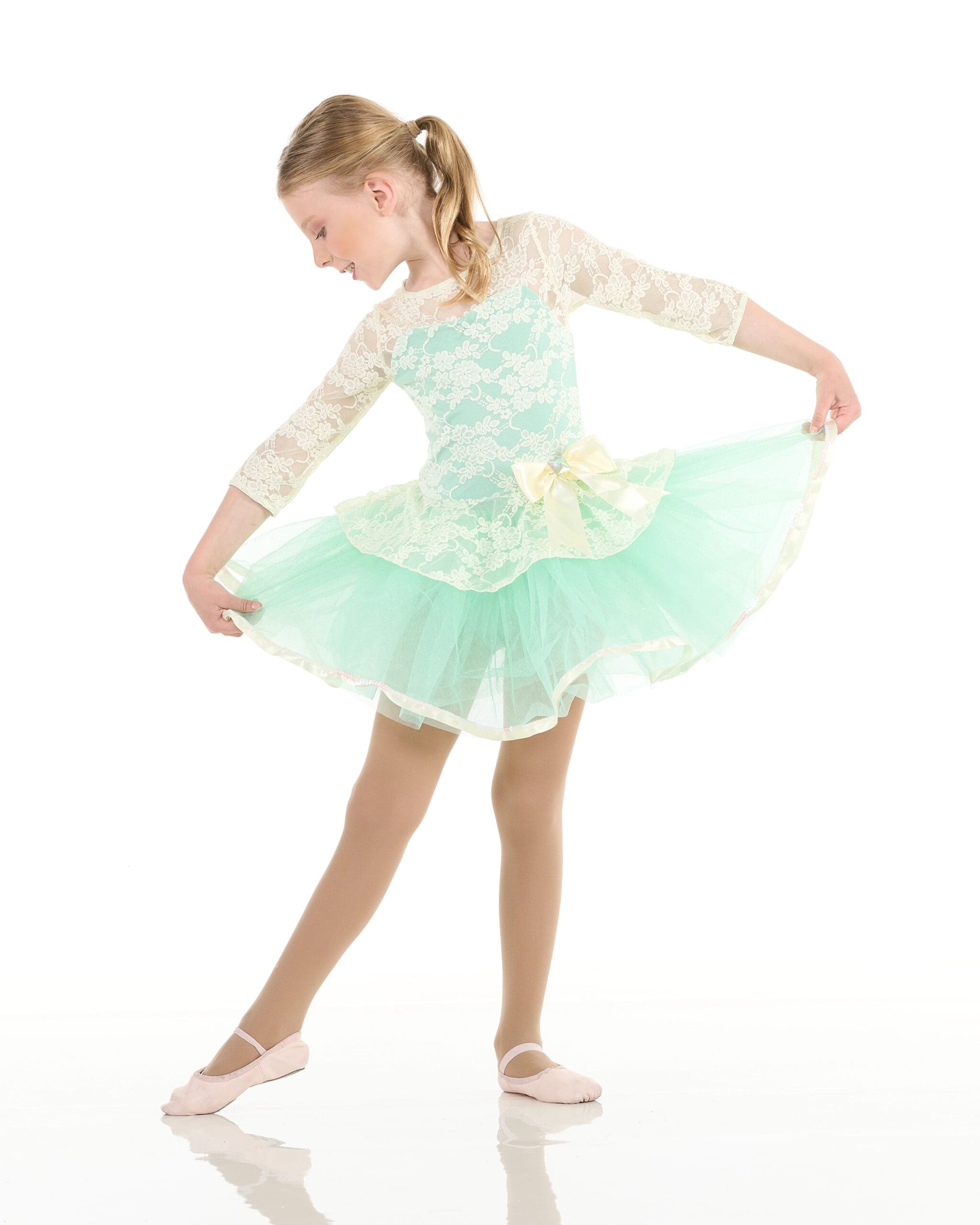 Young girl in a light green and white lace ballet costume with a tulle skirt and bow, standing on one foot, holding her skirt and looking down, posed gracefully against a white background.