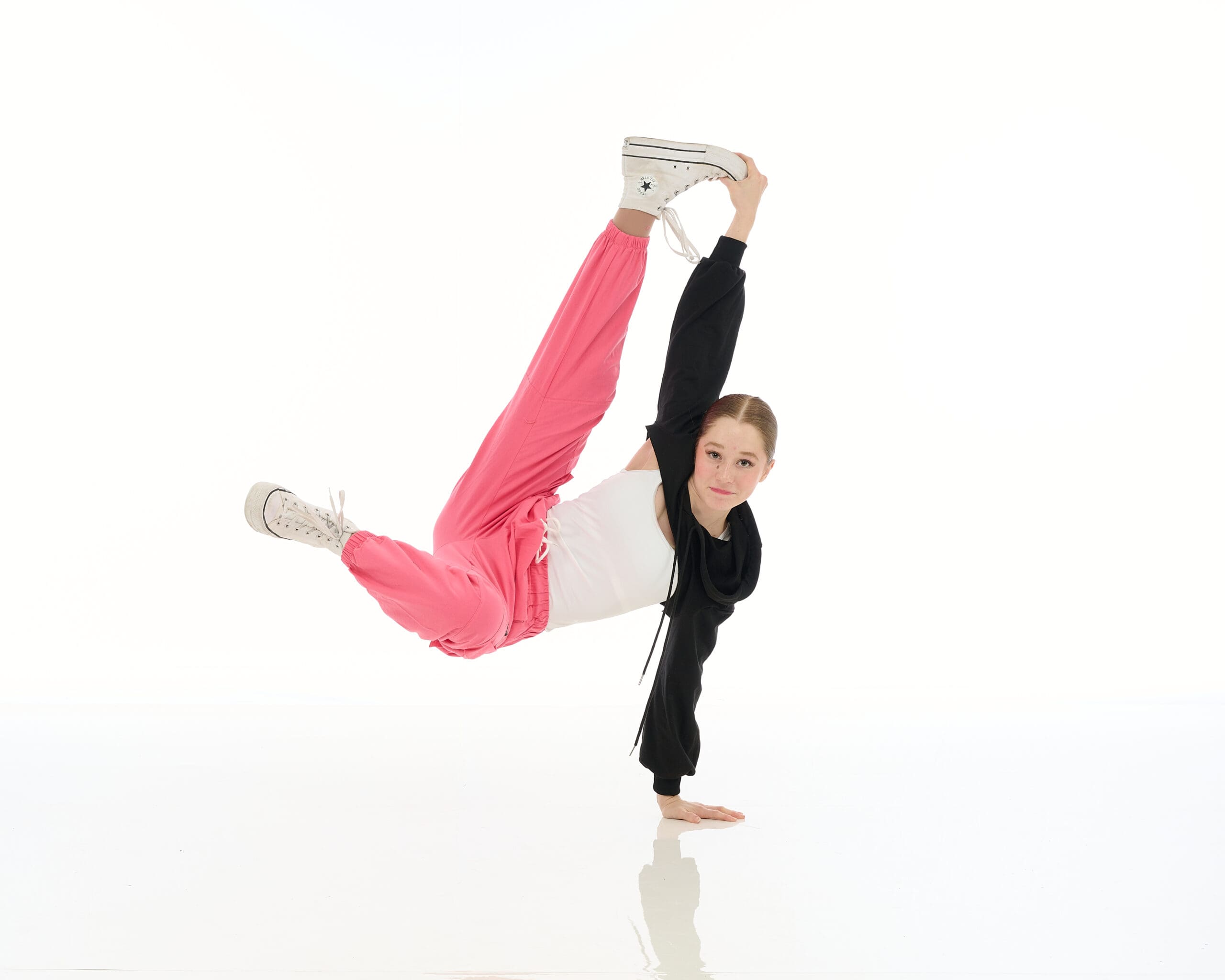 A person in pink pants and a black-and-white top performs a one-handed handstand, holding one leg extended high while balancing, against a white background.