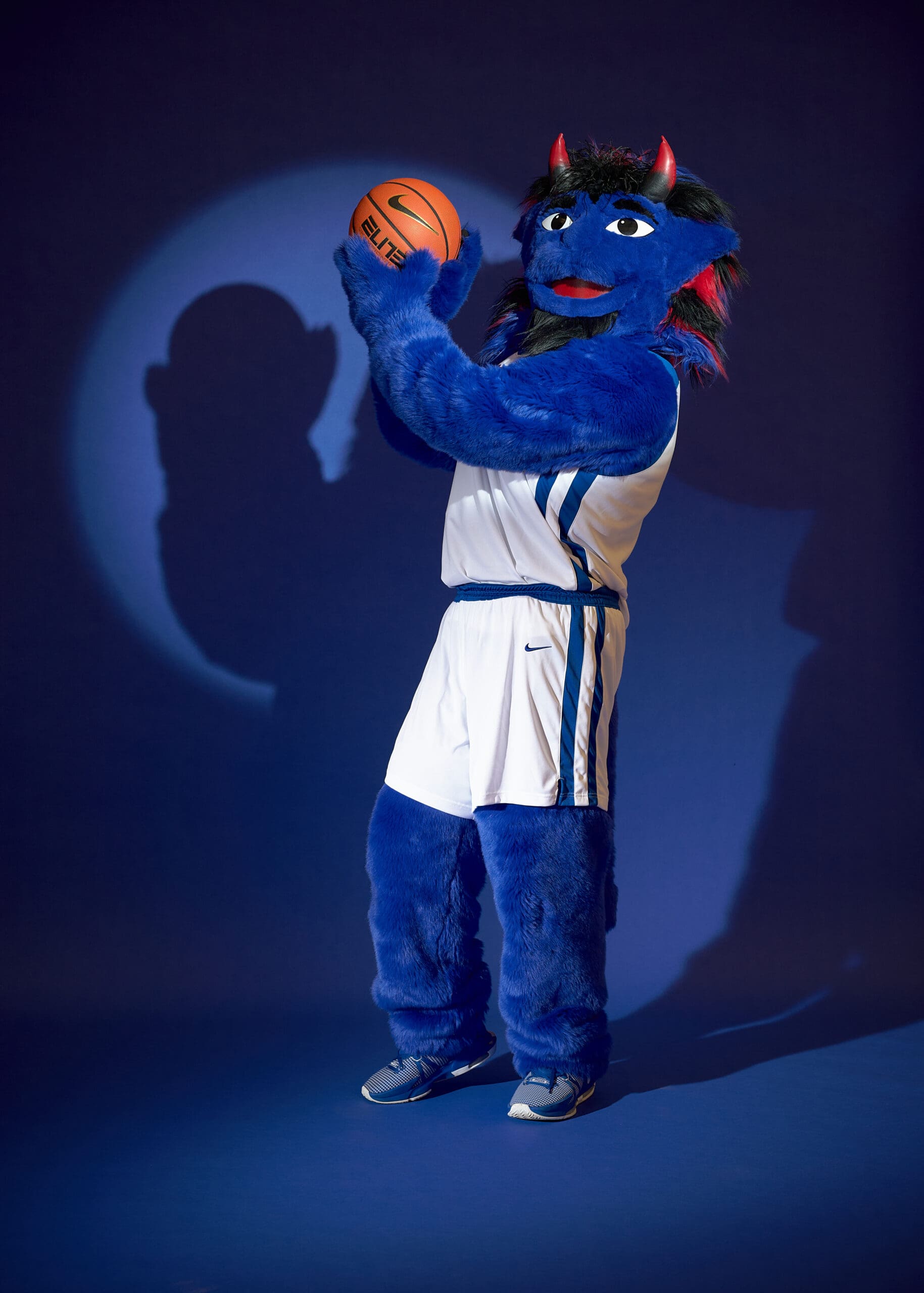 A blue buffalo mascot with red horns, wearing a white basketball uniform and gray sneakers, holds a basketball in front of a blue backdrop, casting a shadow on the wall.