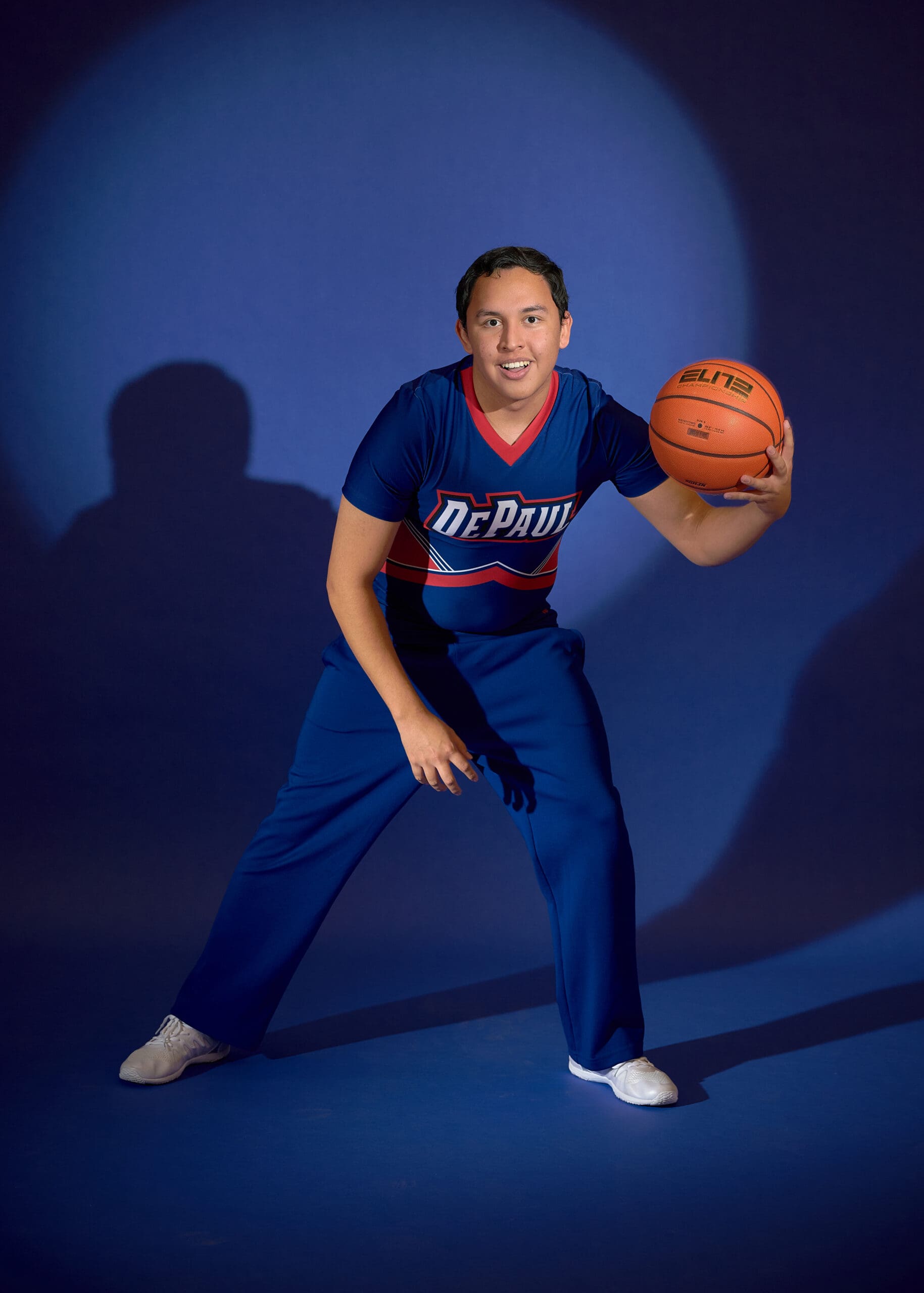 A person in a DePaul cheerleading uniform poses with a basketball, standing on a blue floor against a blue background, smiling and looking at the camera.