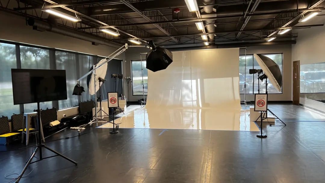 A professional photography studio with large softbox lights, backdrops, tripods, and a white seamless floor, surrounded by windows and lighting equipment. No people are present in the space.