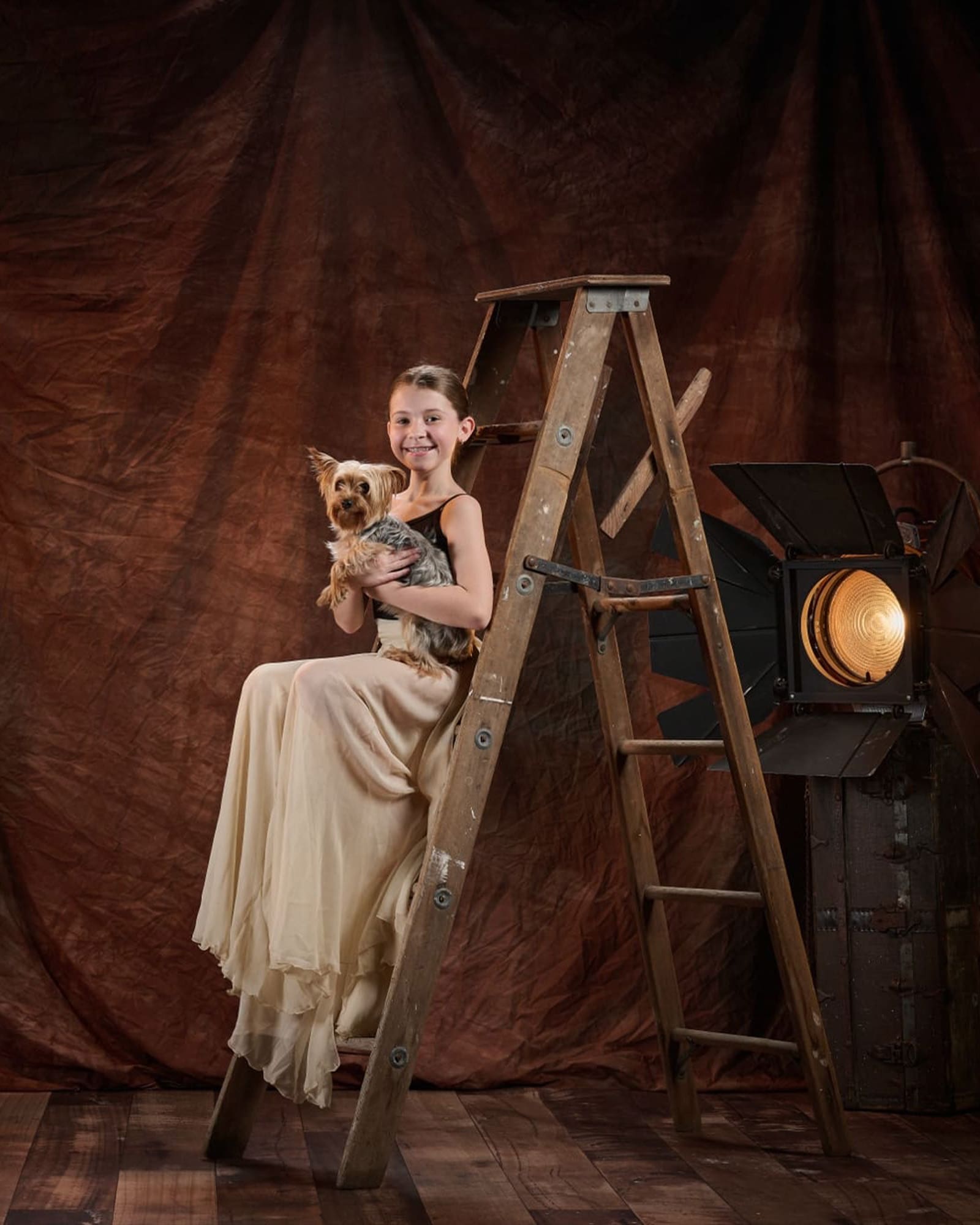 A young girl in a long cream dress sits on a wooden ladder, smiling and holding a small dog. A large studio light is positioned beside her against a brown fabric backdrop.